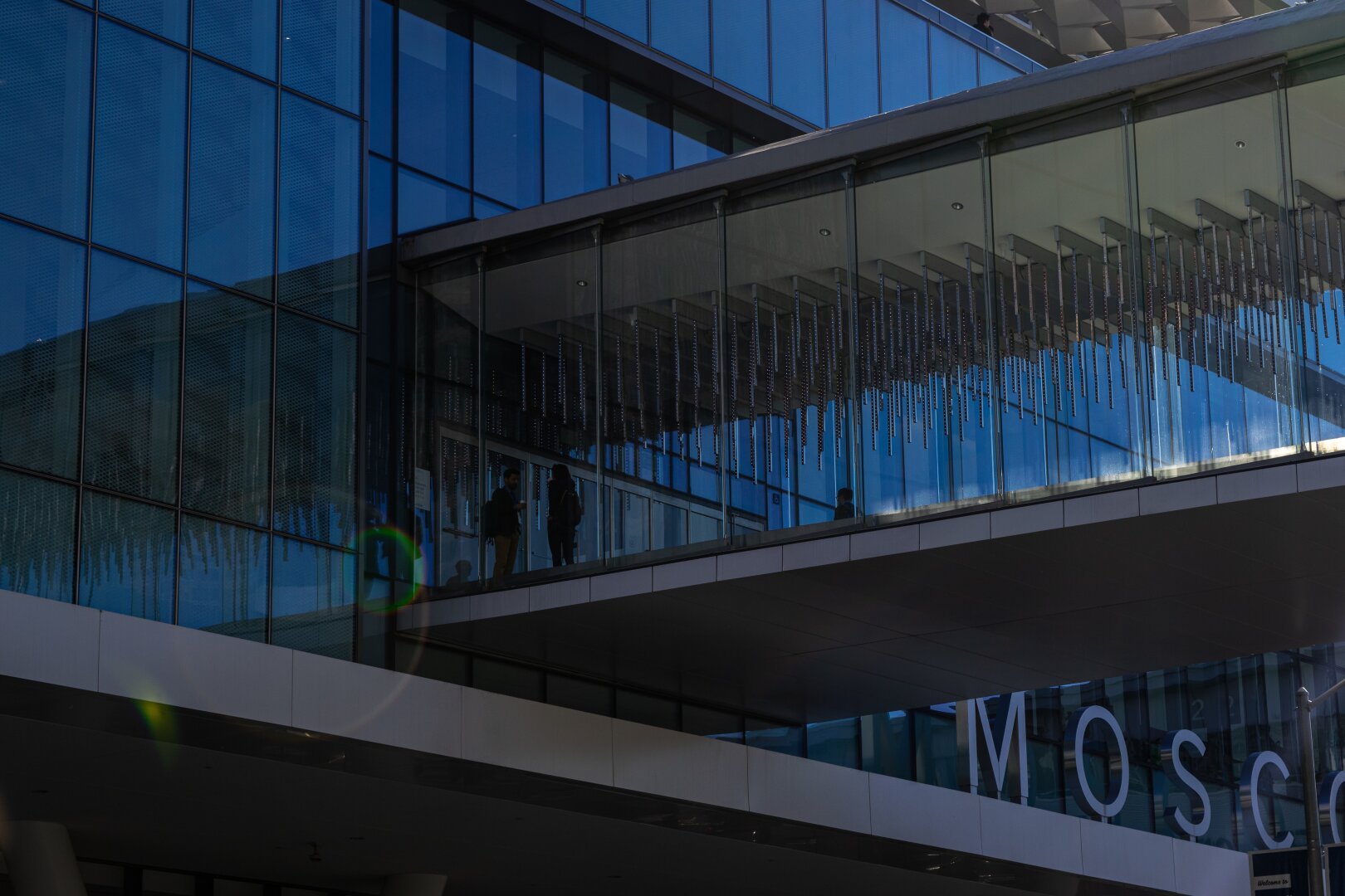 Image of the exterior of Moscone Center in San Francisco, California, on a clear morning. We see one face of the center that has a bridge, walled with glass, connecting the North and South buildings, passing from left to right in the image. Inside the bridge, two silhouettes are mid-discussion, probably planning their day ahead for the conference that is about to begin.