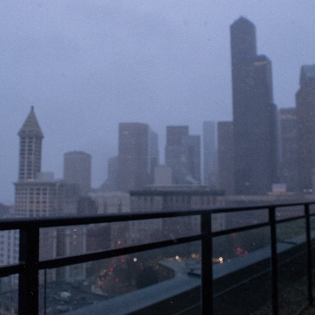 Snowy, late afternoon image of downtown Seattle. The city skyline is blurred with the balcony guardrail in the foreground in hazier focus.