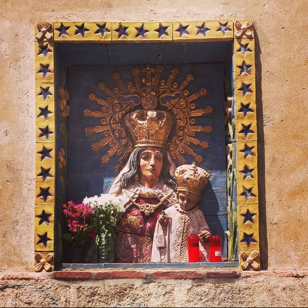 Talla de la fachada exterior en la parroquia de San Sebastián de la Virgen del Carmen coronada Patrona de las Huertas. Se pueden apreciar las ofrendas florales y velas 
La virgen está enmarcada por una cenefa de estrellas de 5 puntas y engalanada con un rico manto, lleva al Niño en brazos también emperifollao y los dos portan sendas coronas de oro que ocupan más de la mitad del espacio
No encontré referencias de quien o cuando se hizo

/Eng/
Carving of the exterior façade in the parish of San Sebastián of the Virgin of Carmen crowned Patroness of the Huertas. You can see the floral offerings and candles 
The virgin is framed by a border of 5-point stars and adorned with a rich mantle, she carries the Child in her arms, also adorned, and both wear gold crowns that occupy more than half of the space.
I found no references to who or when it was done.