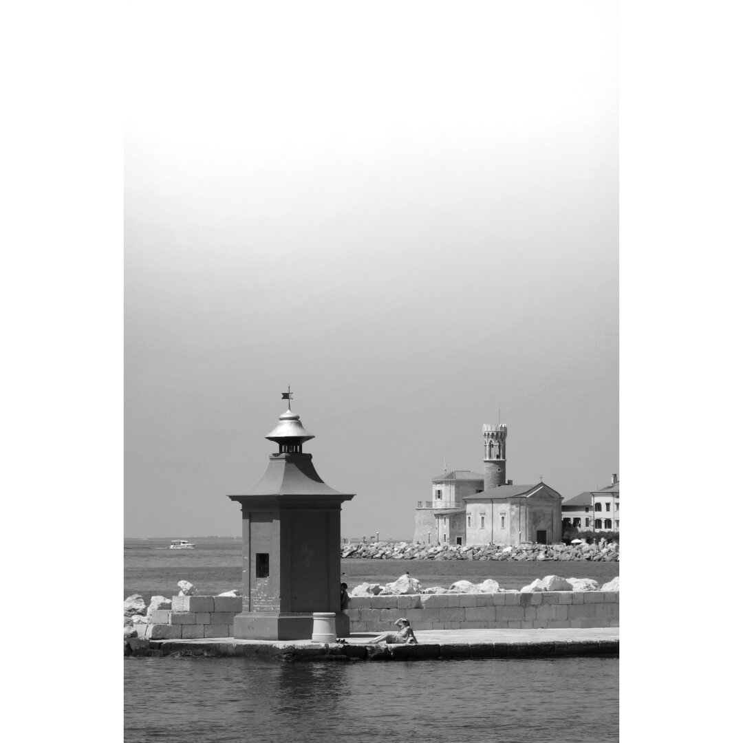 Black and white picture taken from the pier of Pira, showing the old lighthouse, with the tower in the background. Somebody is standing under the shade of the lighthouse, while another person is sunbathing. The sky is clear and the sea is completely calm.