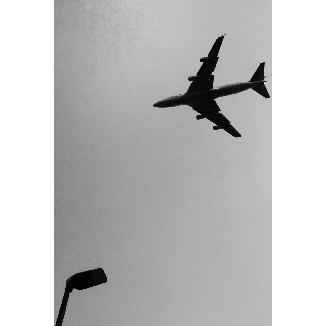 Black and white picture showing a plane, on the top right corner, and a light post, on the bottom left corner. Both elements have a strong contrast against the clear sky, with absolutely nothing else in the picture.