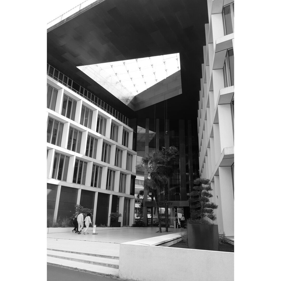 Bnw picture of people entering a modern building, whose roof has openings in the form of trapeze letting in the light.