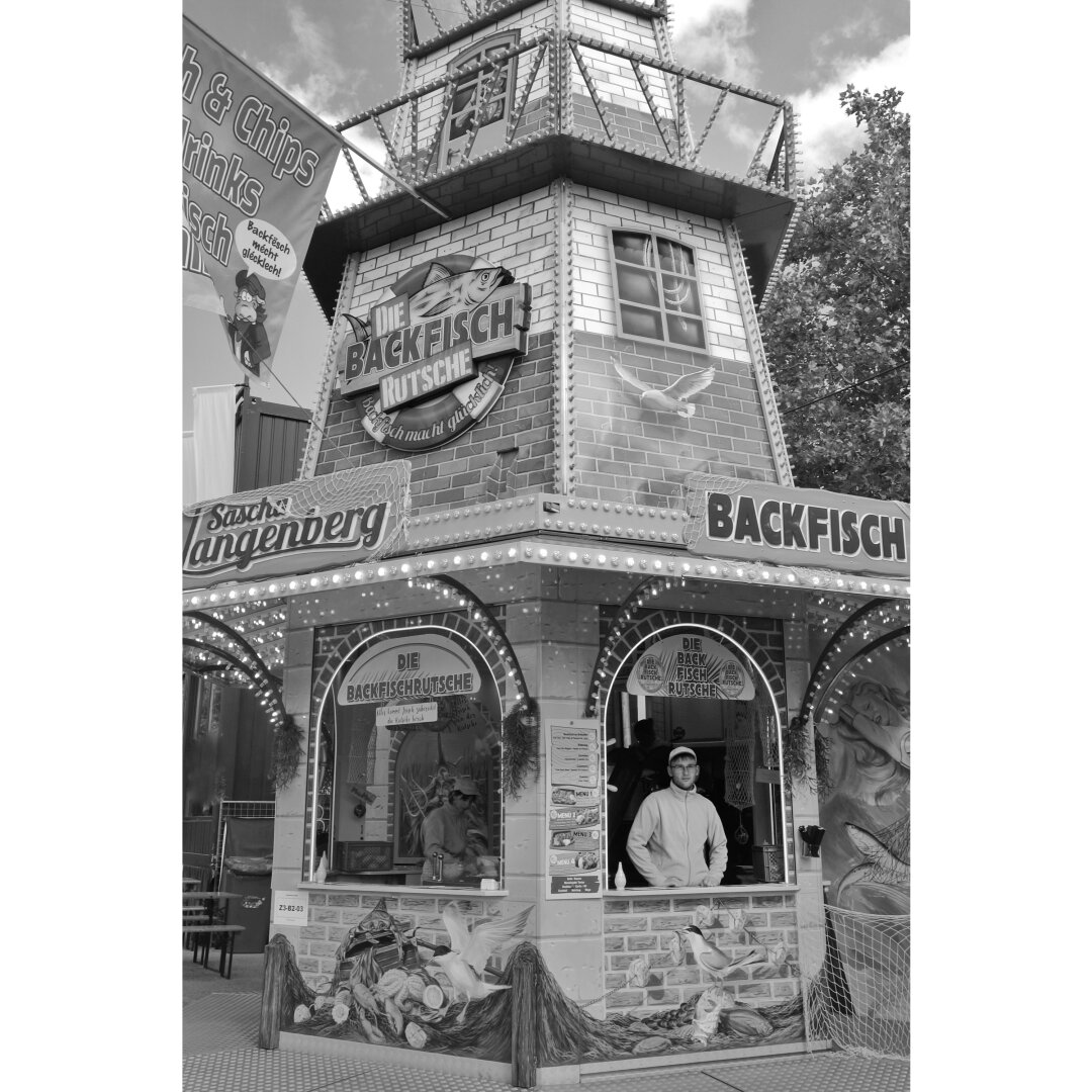 Black and white street photography taken in Luxembourg city during the Schueberfouer showing a man inside a food booth in the shape of a lighthouse selling fish and chips. Backfish is written on a sign above him.