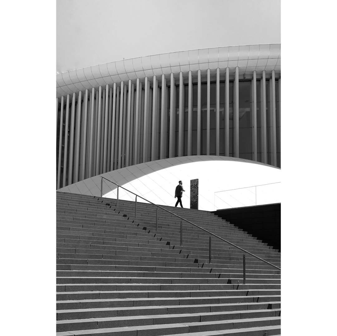 Black and white street photography taken in Luxembourg showing the silhouette of a man approaching the vertical sign on top of the stairs leleading to the Philharmonie.