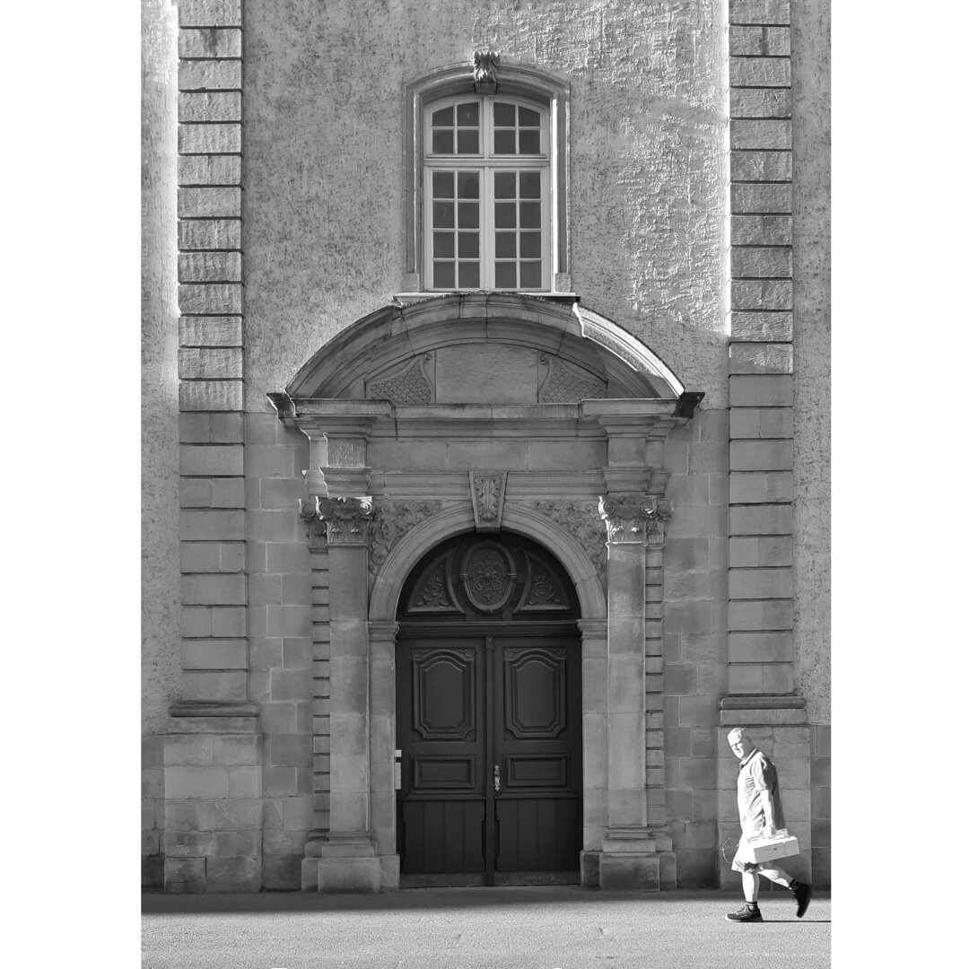 Black and white street photography taken in Luxembourg city showing a man in the sunlight about to pass by the wooden door of the ancient Athenee.