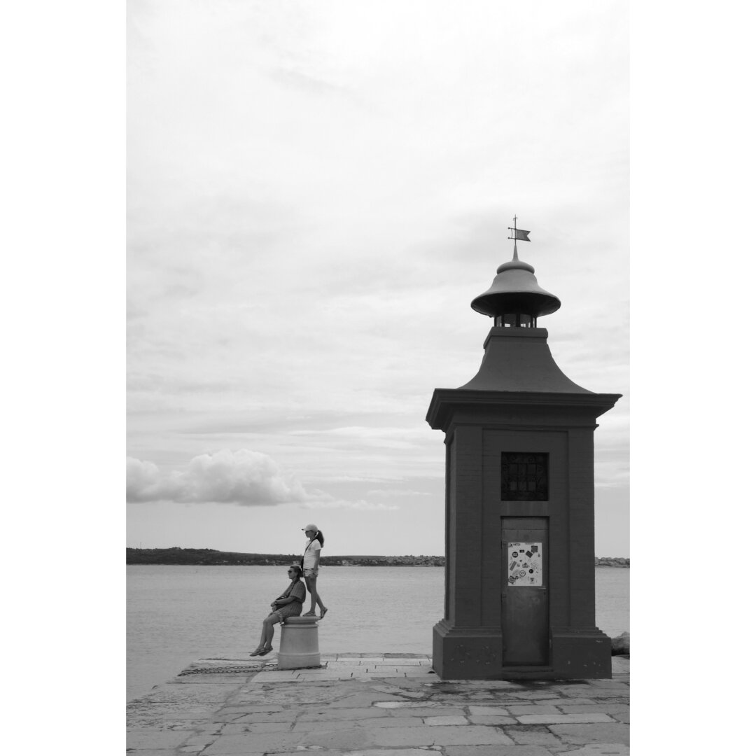 Black and white picture of a woman and a girl sat next to a small lighthouse at the entrance of Piran seafront.