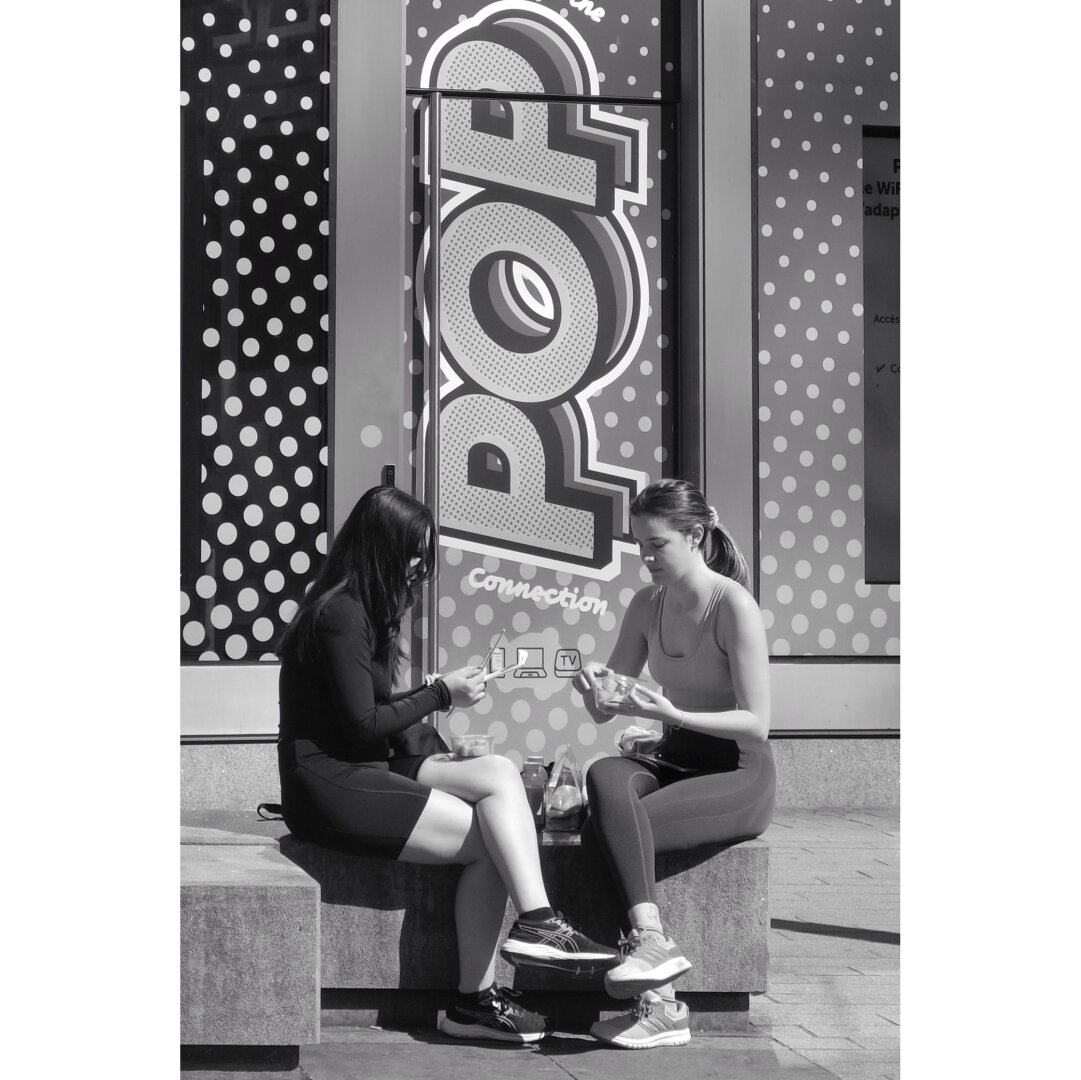 Black and white street photography taken in Luxembourg city showing two girls eating a snack. They sit on concrete blocks. A "Pop" sign decorates a window behind them.