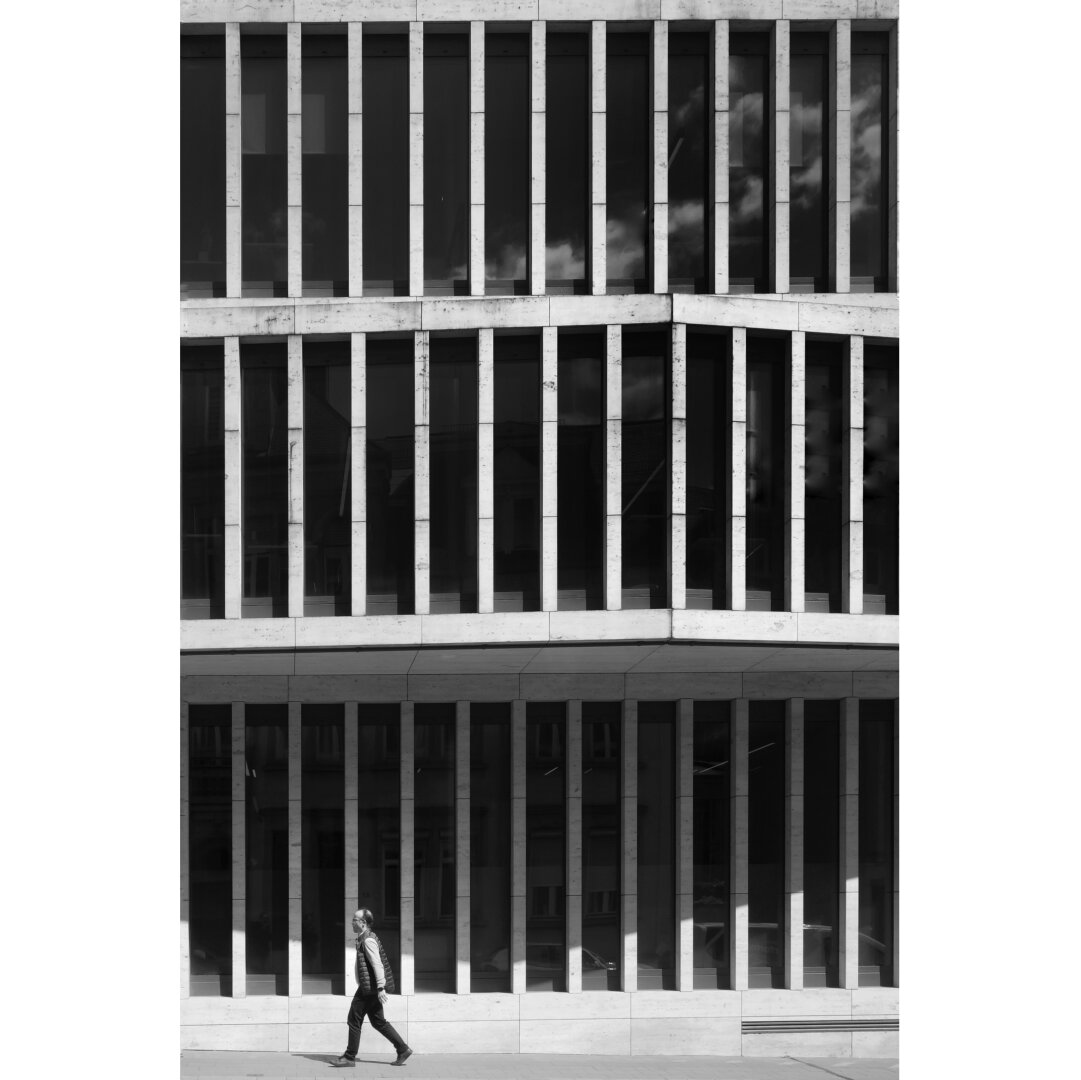 Black and white street photography taken in Luxembourg showing a man walking by the blablack and white facade of a momodern building. The facade is composed of three ranges of vertical windows appeappearing as dark rectangles.  A shadow created by the second range of windows built as an overhang draws a triangle next to the pedestrian.