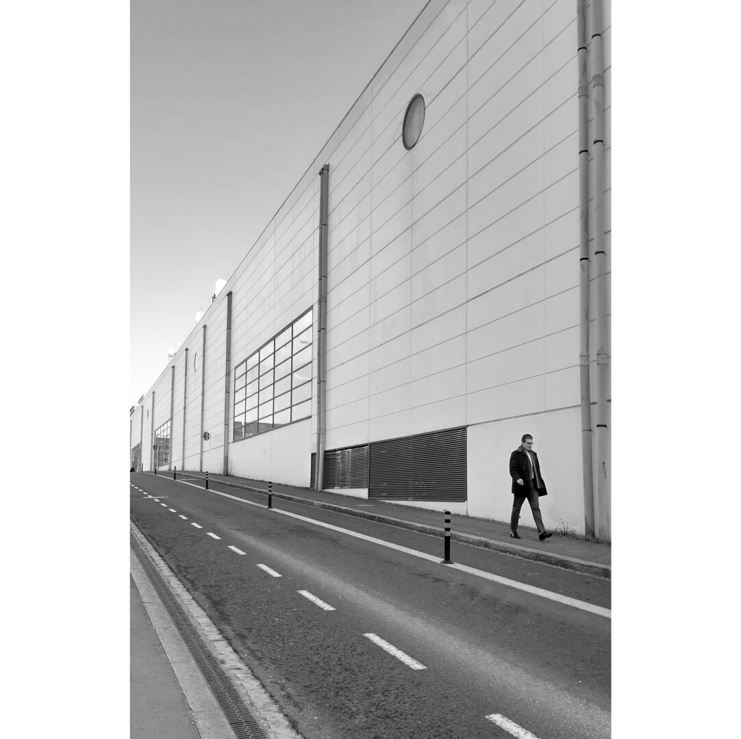 BlBlack and white street photography taken in Luxembourg city showing a man walking in a desert street. He passes by the back of a theater,  composed of blind facades, with only a small circular window near the top.