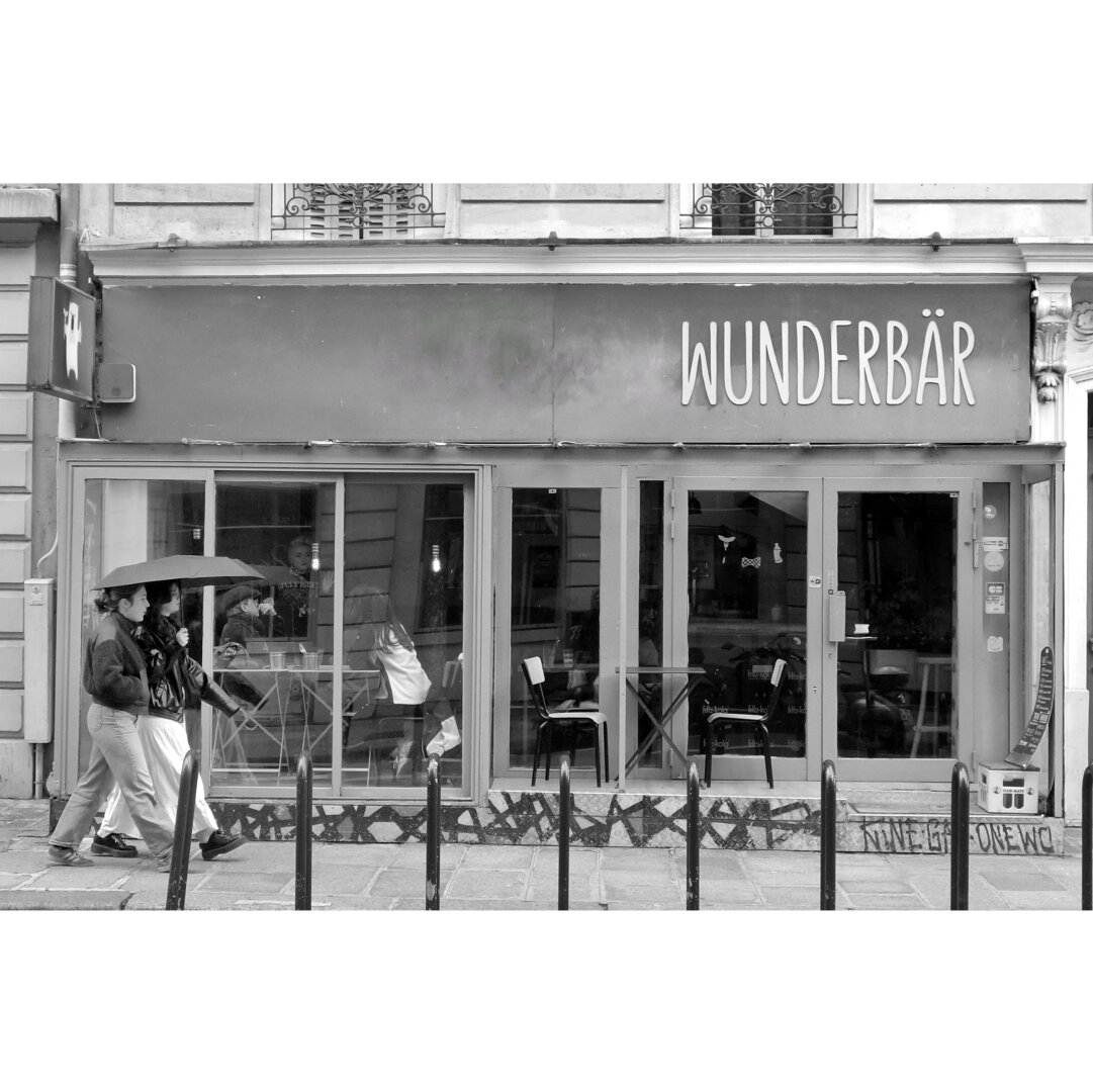Black and white street photography taken in Paris showing two young women under an umbrella walking next to a bar cacalled "Wunderbär" (german pun on wonderful, bear and bar)