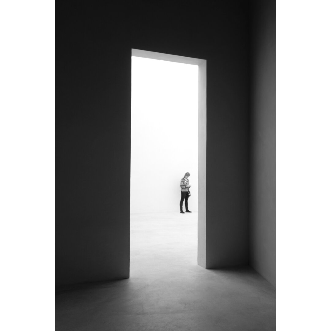 Black and white minimalistic picture taken in the kolomba museum, showing somebody standing in a white room seen through a rectangular opening in a black wall.