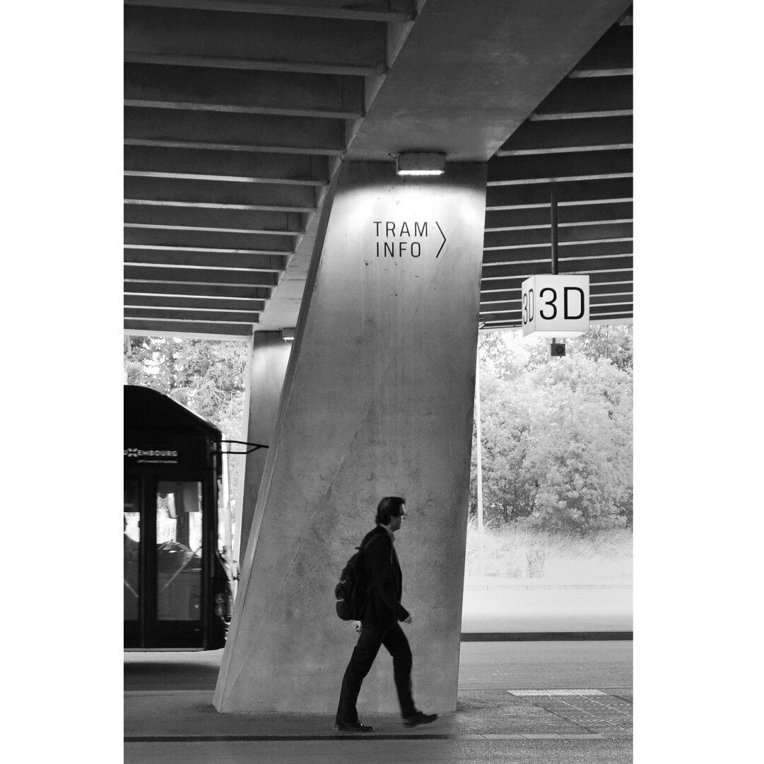 Black and white street photography taken in the bus station of Luxexpo, showing a man walking next to a pillar on which 'tram info' is written. On his left the front part of a bus is parked before the pillar. On his righright a sign with a track number '3D' hangs from the concrete ceiling.