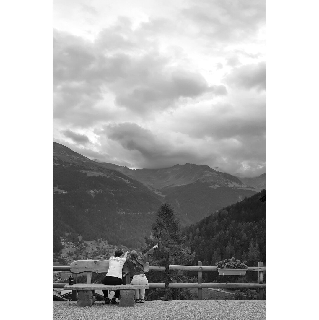 Black and white street photography of two persons sitting on a bench in the village of Grimentz. They are on the bottom left corner of the frame and point their finger to the right but we can only see a cloudy sky and mountains covered with pine trees in front of them. On the right a pot of geraniums hangs on a barrier.