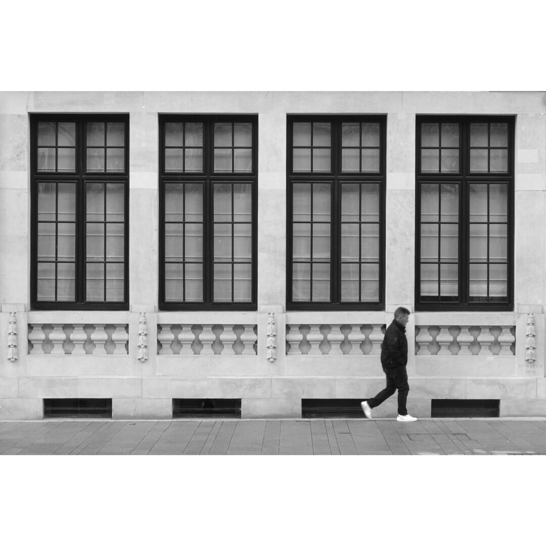 Black and white street photography taken in Luxembourg city showing a man walking on the pavement under a range of four rectangular windows.