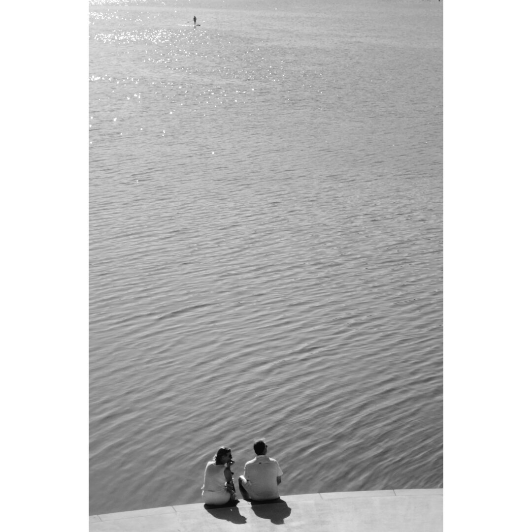 Black and white picture of a couple on the edge of the Stausee, seen from the back. In the distance a tiny silhouette can be seen paddling on the lake.
