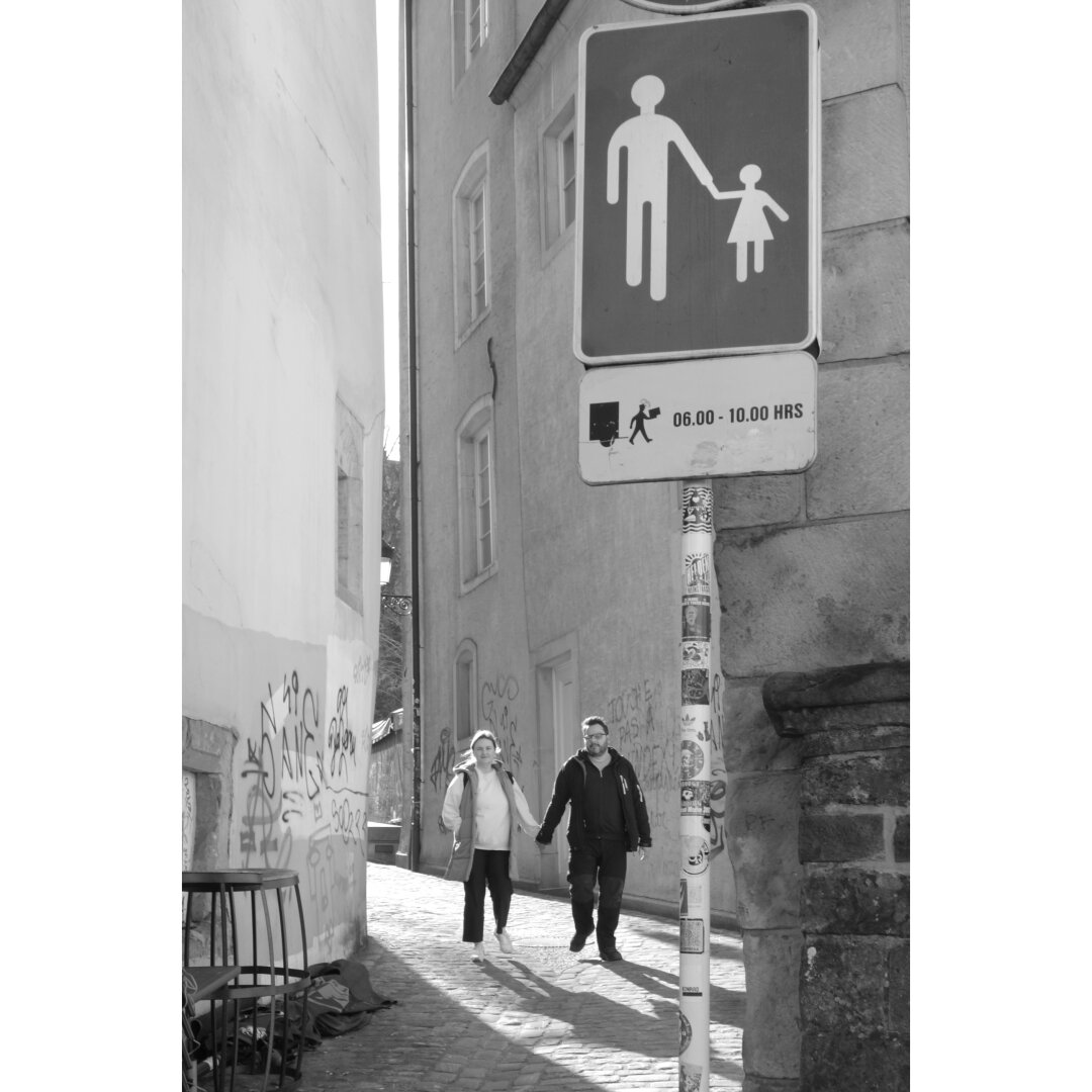 Black and white streetphotography taken in Luxembourg city showing a couple walking down a narrow pedestrian street. They hold their hands, thus respecting the road sign in the foreground showing an adult and a child holding their hands.