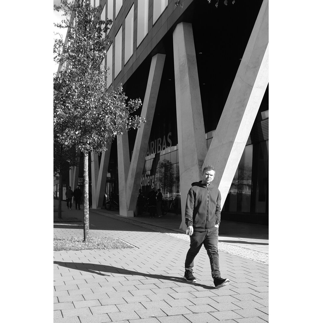 Black and white street photography taken in Luxembourg city showing a man approaching. Behind him, the ground floor of a building is made of concrete pillars drawing a W.