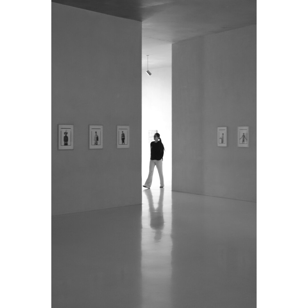 Black and white photography taken in a room of the Kolumba museum, showing a woman standing in the light of the opening created to exit a rather dark exhibition room, on the walls of which a few pictures are displayed. The silhouette of the woman and of the bright opening is reflected by the floor.