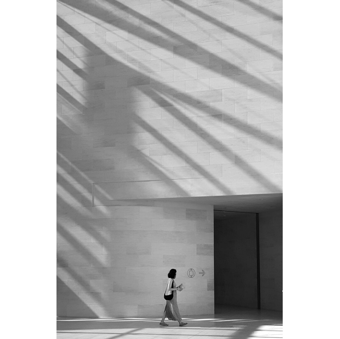 Black and white picture of a woman looking tiny against the huge wall at the entrance of the Mudam. On the right, a black square is the entrance of a darker room. Behind her and on the upper part, the shadows create a pattern of darker lines on the wall.