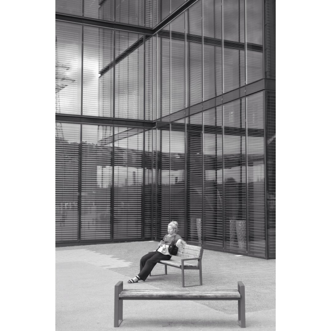 Black and white street photography taken in Luxembourg city showing a woman sitting on a bench next to a corner creating reflections of the glass facade behind her. In the foreground slightly on the right, there is another bench, empty.