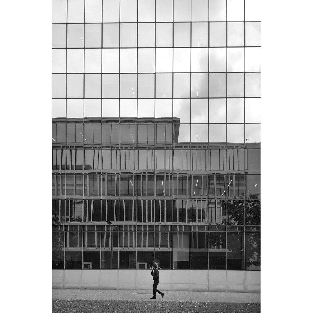 Black and white street photography taken in Luxembourg city showing somebody passing by a facade made of glass reflecting another building.