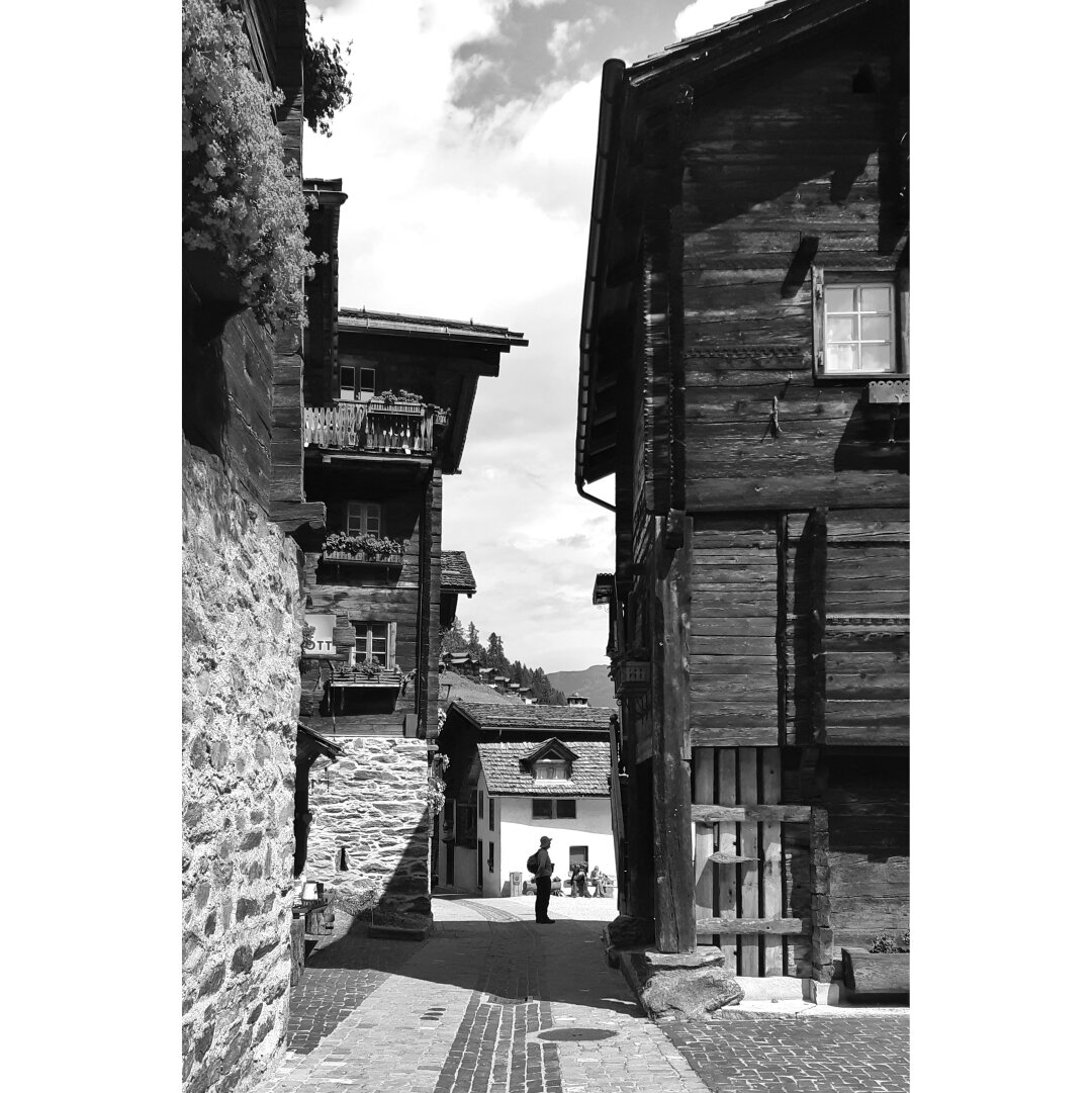 Black and white picture taken in a narrow street of Grimentz village showing the silhouette of a man between traditional wooden houses.