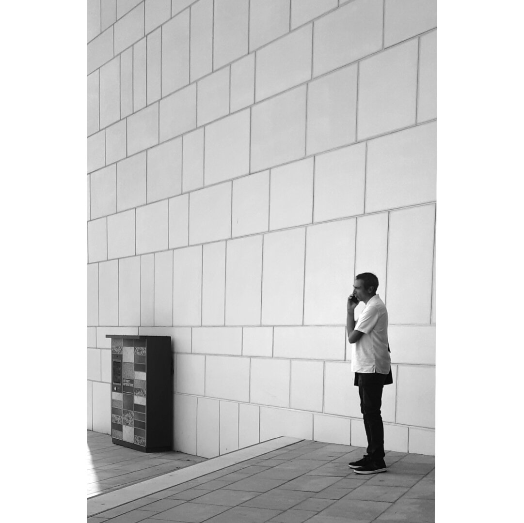 Black and white street photography taken in Luxembourg city showing a man standing at the wall of the national Library with his phone next to his ear. In front of him, next to the white wall, there is a self service box to collect books ordered from the website.