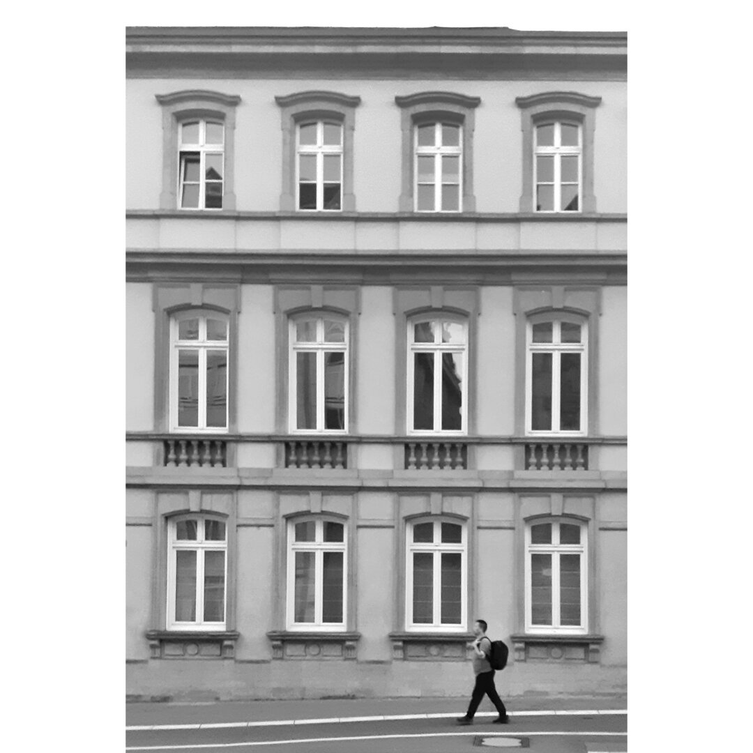 Black and white street photography taken in Luxembourg city showing a man walking on the Boulevard Roosevelt. He passes by the hotel des terres rouges, and its facade with twelve windows.