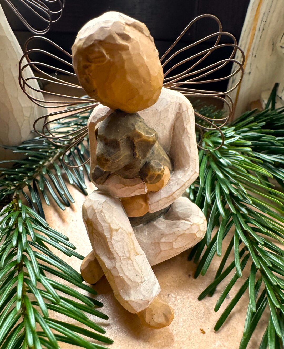 Ein kleiner Engel mit Drahtflügeln sitzt zwischen Tannengrün auf einer Fensterbank und hält einen kleinen Hund im Arm.