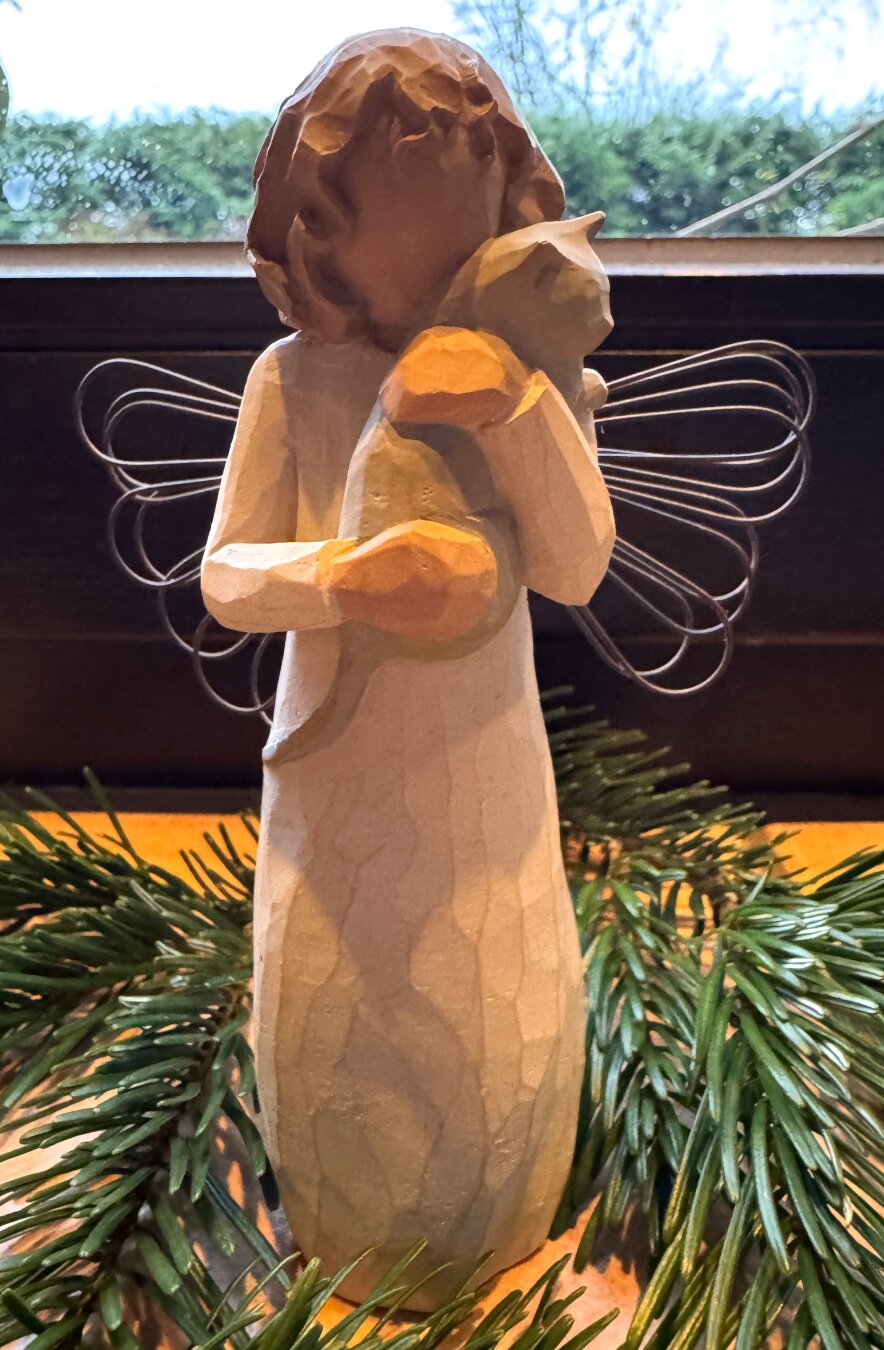 Ein auf einer Fensterbank im Tannengrün stehender Engel mit Drahtflügeln hält liebevoll eine Katze im Arm.