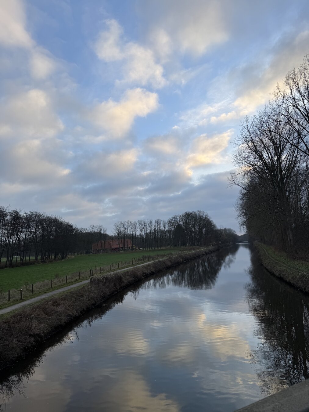 Ein Fluss, die Ems, umgeben von Bäumen, Sträuchern und Uferwegen, blauer Himmel mit leichter Bewölkung, die sich im Wasser spiegelt