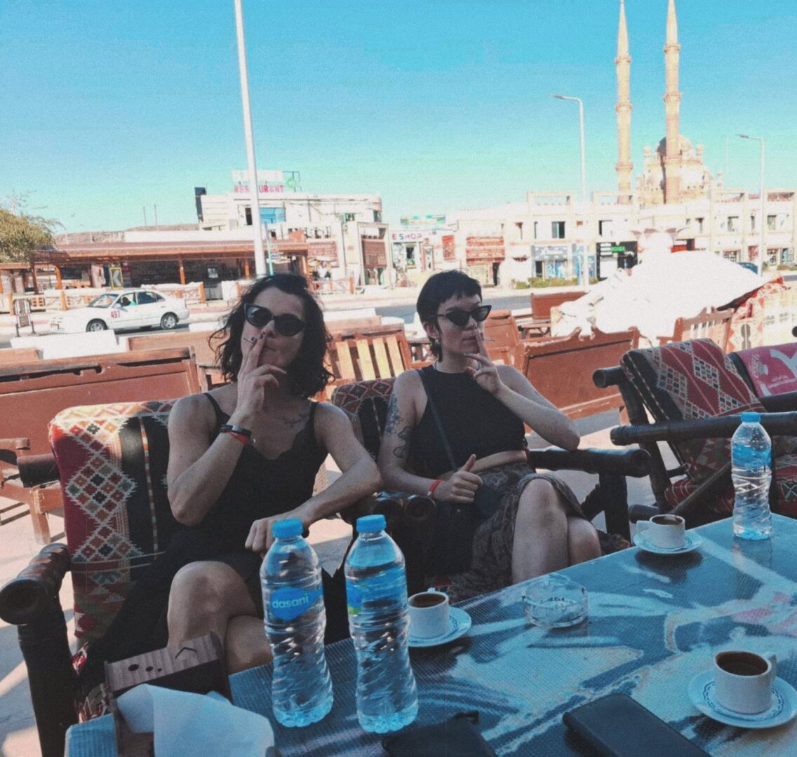 two women smoking with 3 cups of turkish coffee in front of them and a mosque behind them