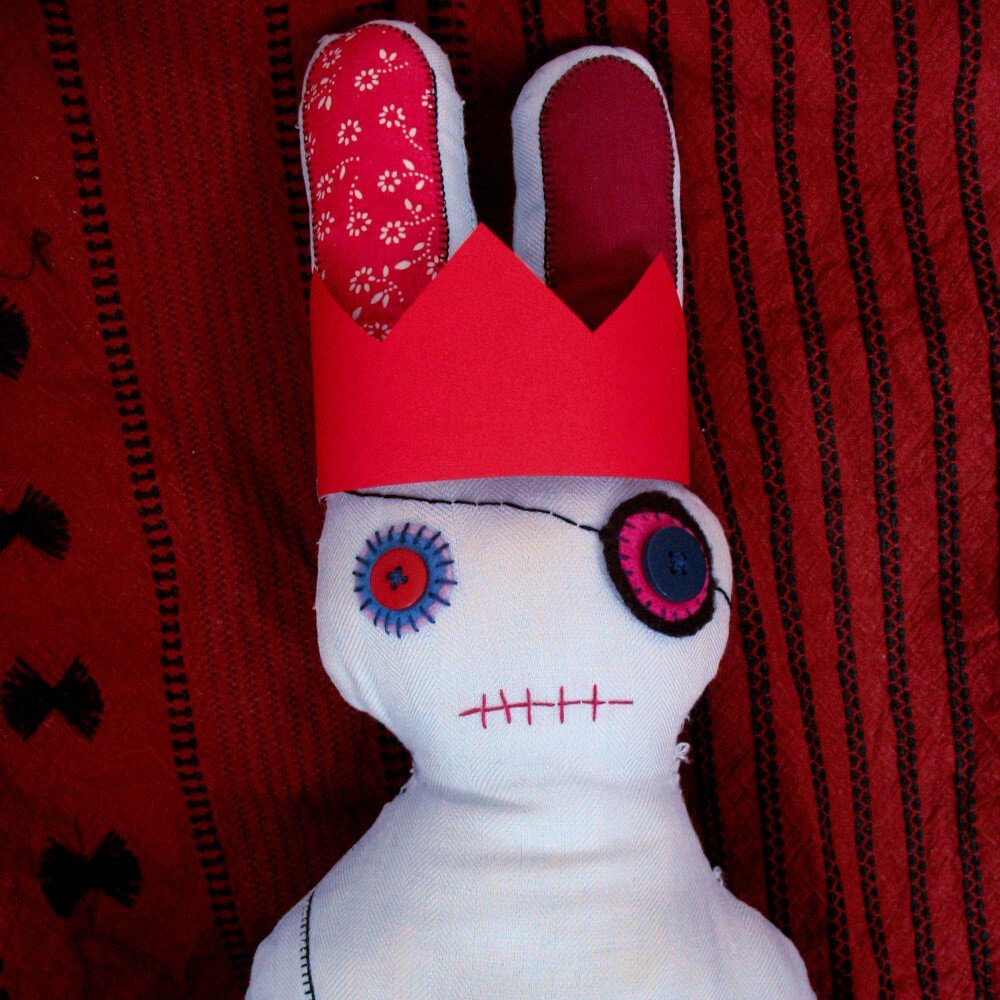 Stuffed bunny doll made with light blue fabric, with buttons as eyes, an eye patch and long red ears, wearing a red cardstock crown.