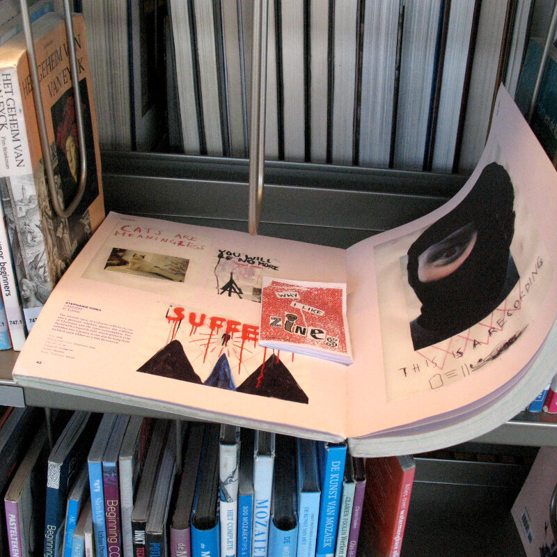 A library book about zines is opened on a library shelf between lots of art books. On top of the opened pages is a mini-zine titled Why I Like Zines with a red collage cover.