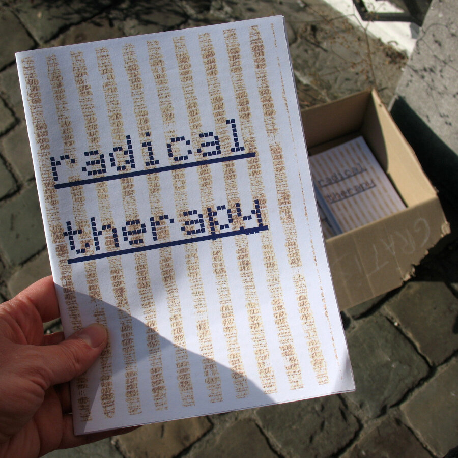 My hand holding a zine titled "Radical Therapy. In the background is a cardboard box on the pavement with the remaining copies of the zine.