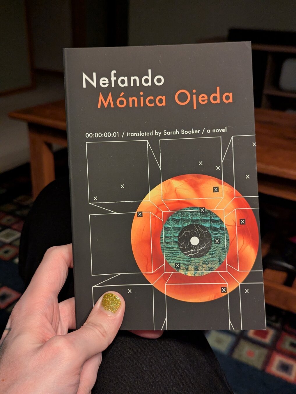 A photo of my hand holding a paperback copy of Nefando by Mónica Ojeda. My thumb nail is painted a shimmery green. The background is blurred, but a coffee table and a rug with a colored square pattern can be seen. The cover of the book is black with a reddish orange orb on it. The color turns to a textured green as it gets closer to the center. Then black with white branching lines. Then just pure white. It resembles an eye. There are several black squares with a white 