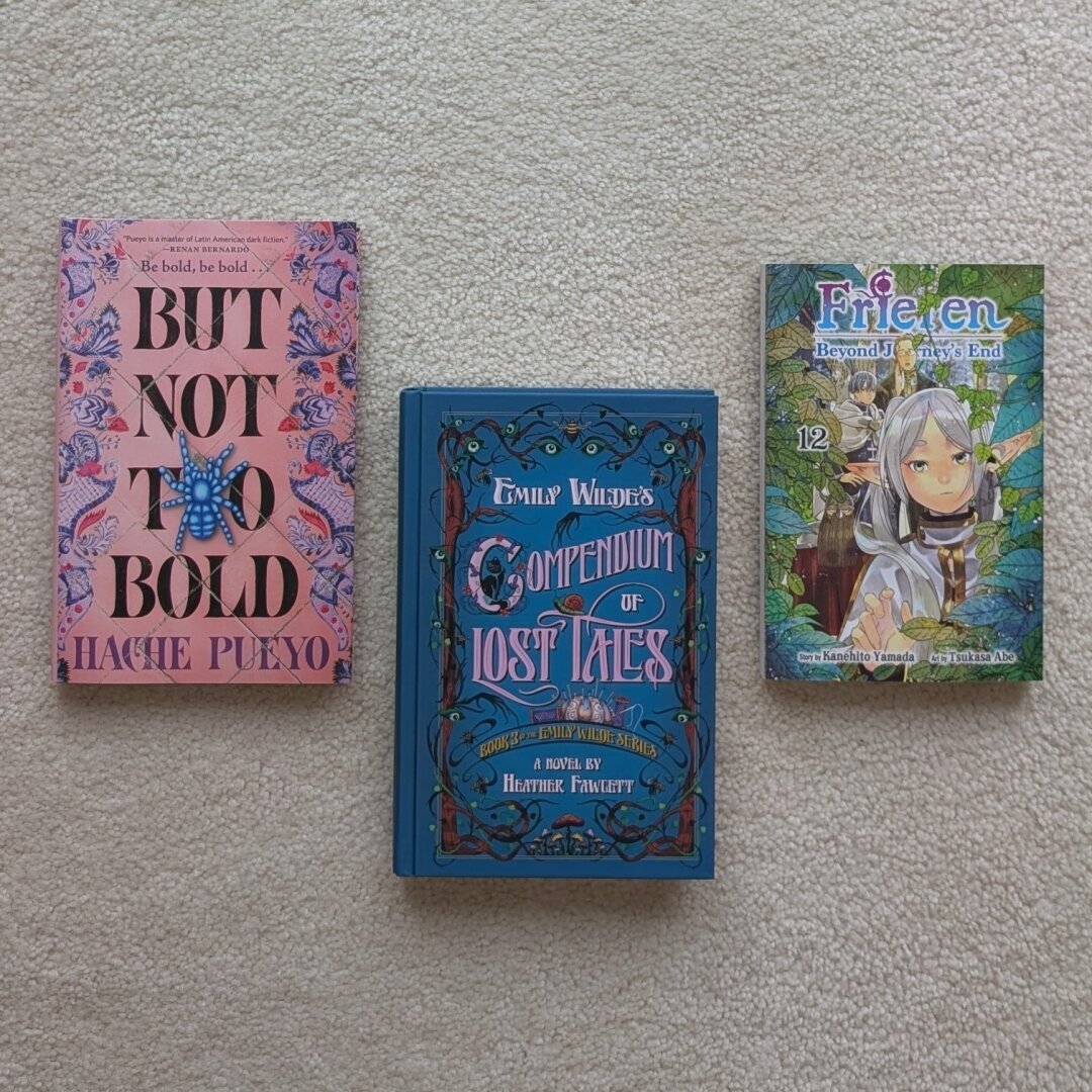 A picture of three books sitting on the carpet face up. They are placed in one row of three with the first and third slightly higher than the middle. The books are:

But Not Too Bold by Hache Pueyo
Emily Wilde