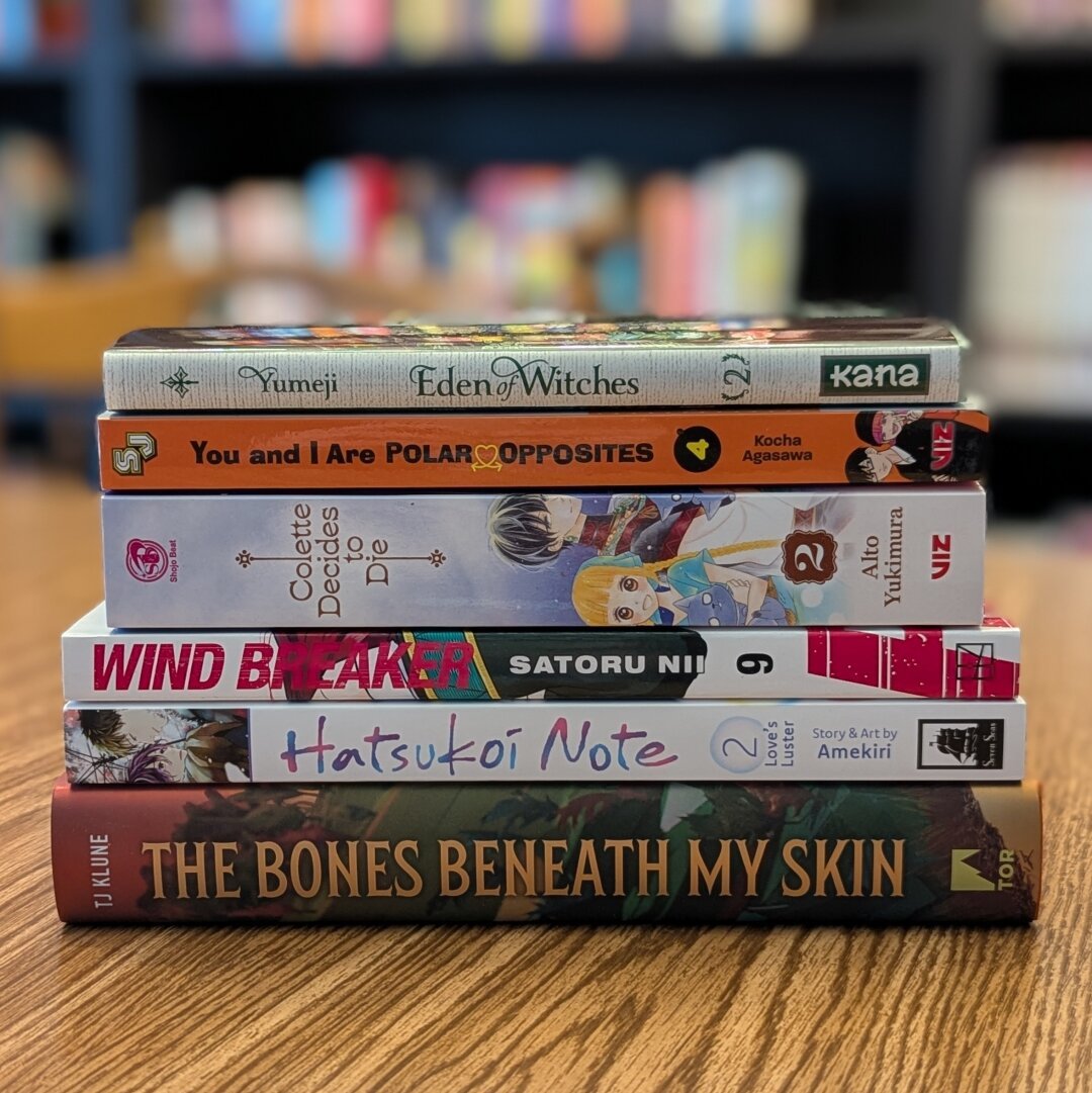 A photograph of six books stacked on top of each other sitting on a table. Their spines are facing out to show their titles. The background is blurred, but bookcases can be seen behind them. The six books from top to bottom are:

Eden of Witches Vol. 2 by Yumeji
You and I Are Polar Opposites Vol. 4 by Kocha Agasawa
Colette Decides to Die Vol. 2 by Alto Yukimura
Wind Breaker Vol. 9 by Satoru Nii
Hatsukoi Note Vol. 2 by Amekiri
The Bones Beneath My Skin by TJ Klune