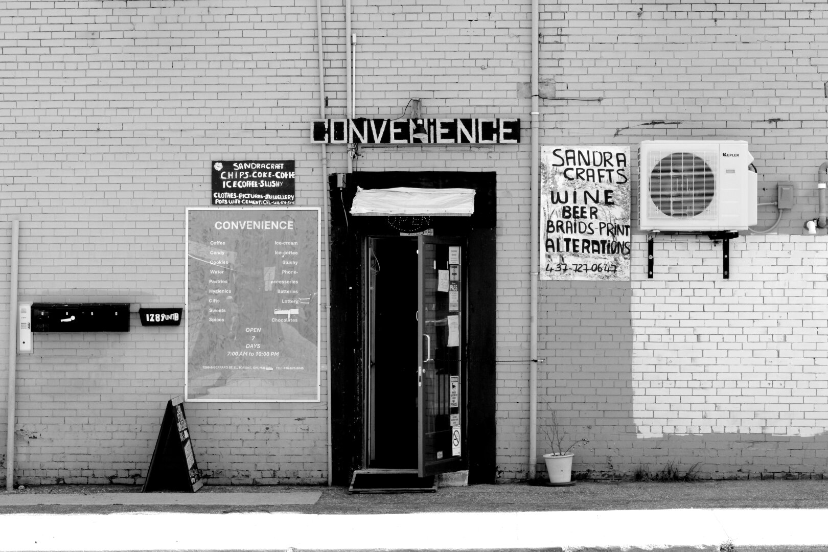 A single door in a brick wall, labelled “Convenience” with a handmade sign. Beside the door, another handmade sign reads, “Sandra Crafts. Wine. Beer. Braids. Print. Alterations” with a phone number.