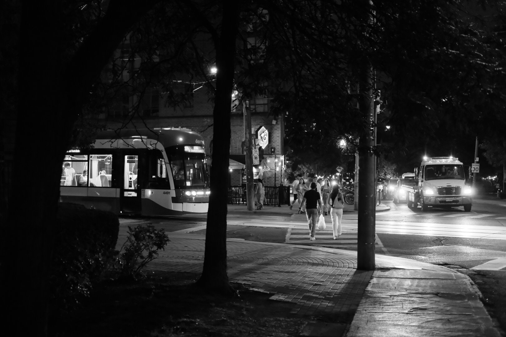 Night. Looking down the street at the lights of the intersection ahead past shadowy trees. There’s a streetcar at left, pedestrians framed by two tree trunks at centre, and the lights of an approaching truck and line of cars at right.
