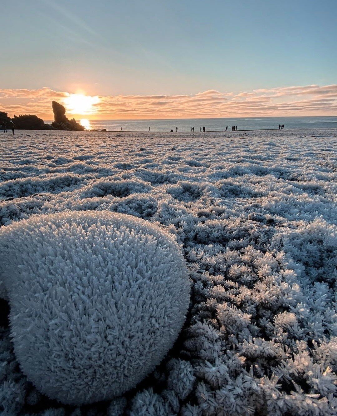 Photography. A color photo of a stretch of coastline in Iceland. The stony landscape is covered with small frosty ice flowers. In the foreground lies an almost heart-shaped large round stone in this cool landscape. The sea and the outlines of people on the beach can be seen in the background. Some large rocks can be seen on the left and a friendly blue sky with veil clouds and orange sunbeams complete this beautiful landscape photo.