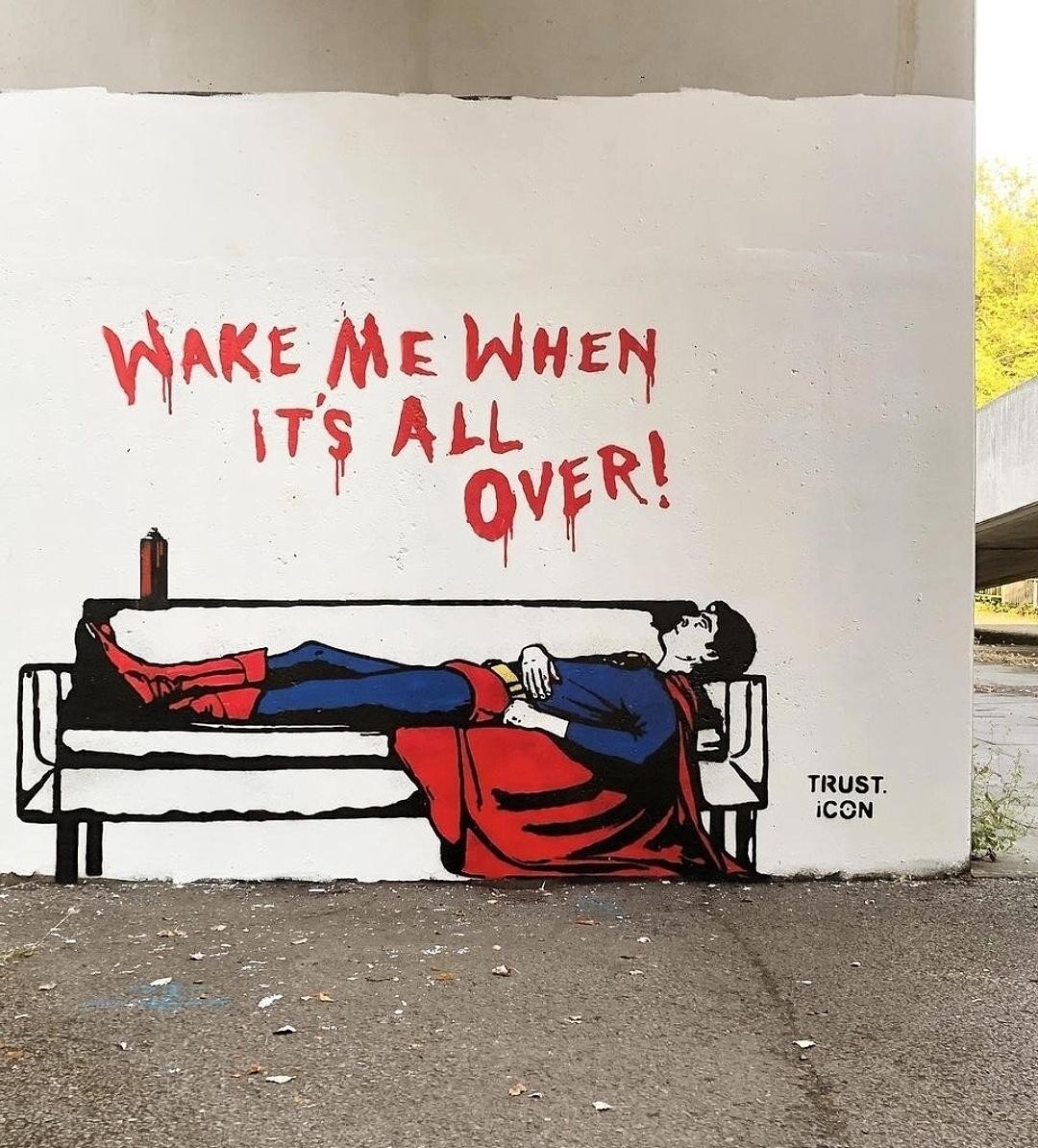 Streetartwall. A funny mural of a very unnerved Superman was sprayed onto a beige-colored exterior wall. The background is painted white. Superman can be seen in his full superhero outfit on a white sofa. There is a spray can of red paint on the sofa and he has sprayed a text in capital letters on the wall above it: 