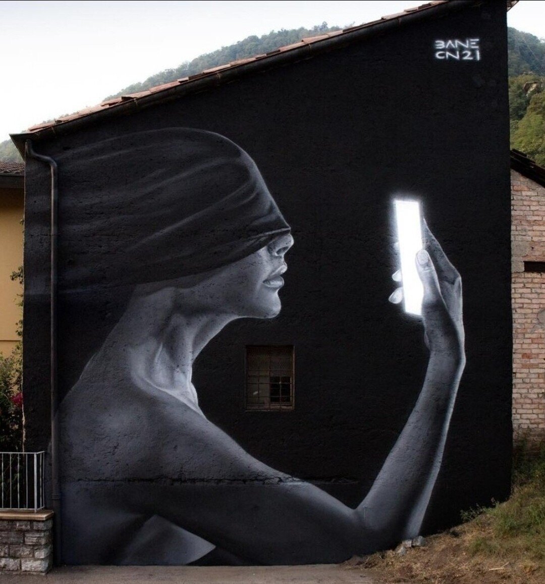 Streetartwall. Small, black-painted and sloping house wall. You can see a mural with the upper body of a young woman sitting in the dark. Her eyes are covered with a black cloth. In her right hand, she holds a glowing white smartphone in front of her face, as if she is trying to recognize something. The very dark mural in gray, black and white creates a fascinating atmosphere of abandonment in a modern society.