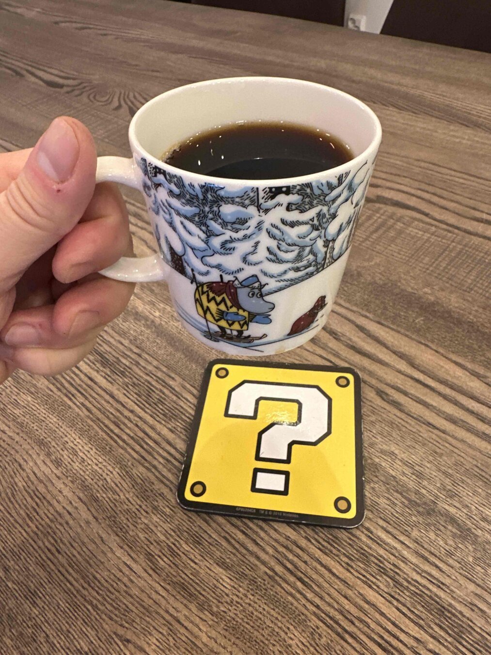 Hand holding an Arabia Moomin coffee cup with black coffee over a Super Mario question block themed coaster on a wooden tabletop.