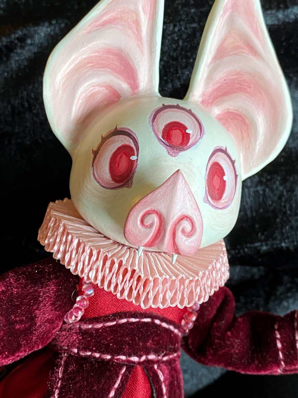 A closeup of a handmade albino bat doll. She has large pointy ears, three red eyes, a pink neck ruff, and a dark red velvet dress.