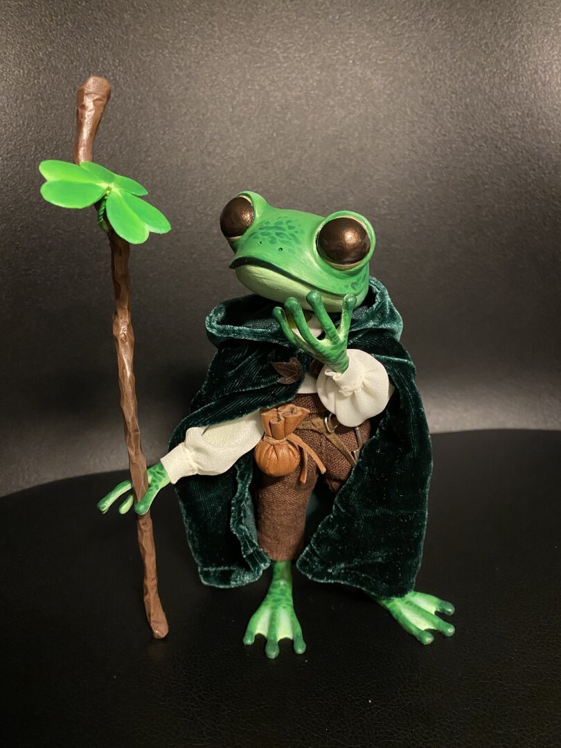 A little handmade frog doll is standing on a black chair. He is painted green with big round black eyes, and is holding a wooden staff with a three leaf clover poking out of it in one hand, while the other hand is helt near his chin. He seems to be looking at the staff in wonder. He is wearing a white flowey shirt, cropped brown pants, so his green feet are showing, and a dark green velvet cape with a hood. On his belt he has a leather pouch and an old metal key.