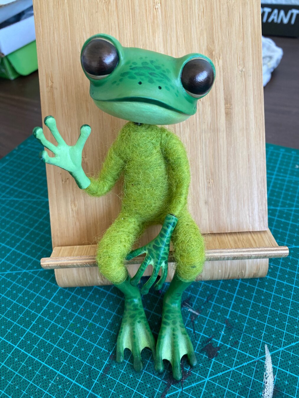 A little green handmade frog doll sitting on a bamboo phone stand. It has green painted head, hands and feet, and a green needlefeltet body. It is posed sitting with one hand between it's legs and one hand up in a wave.