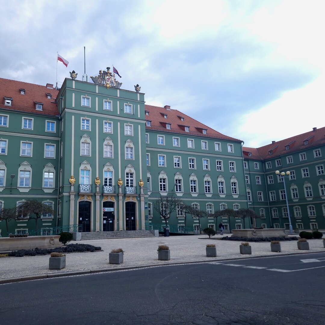 Large, green building with white and gold decors and red roof. Building servers as the City Hall of Szczecin. On top of the building there's a flag of Poland and Szczecin.