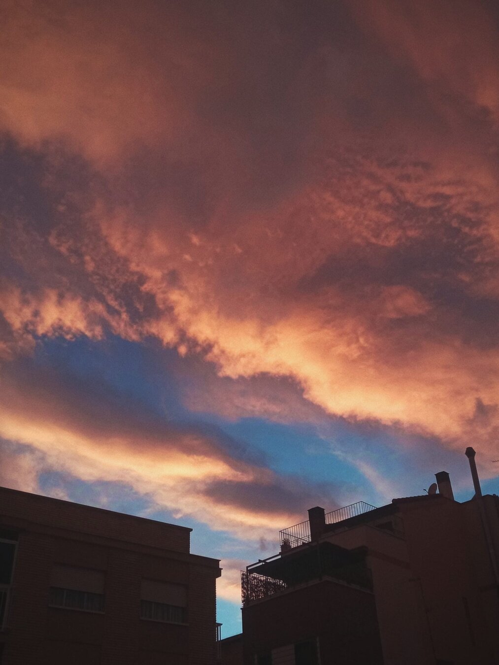 Fotografía del cielo en un atardecer en la ciudad de Murcia. En la parte inferior de la foto se ven fragmentos de siluetas de edificios. El cielo azul está cubierto por nubes de tonos anaranjados, rosados y violáceos.