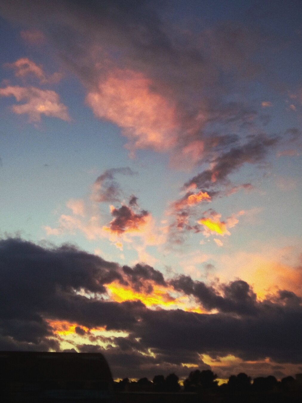 Una puesta de sol con nubes de tonos oscuros y rojizos. Las nubes más bajas son oscuras, casi negras, mientras que las nubes más altas muestran tonos anaranjados y rosados, con destellos amarillos cerca del horizonte. Se ve una silueta de árboles y posiblemente un edificio en la parte inferior de la imagen. El cielo es un degradado de azul oscuro a tonos más claros cerca del horizonte.